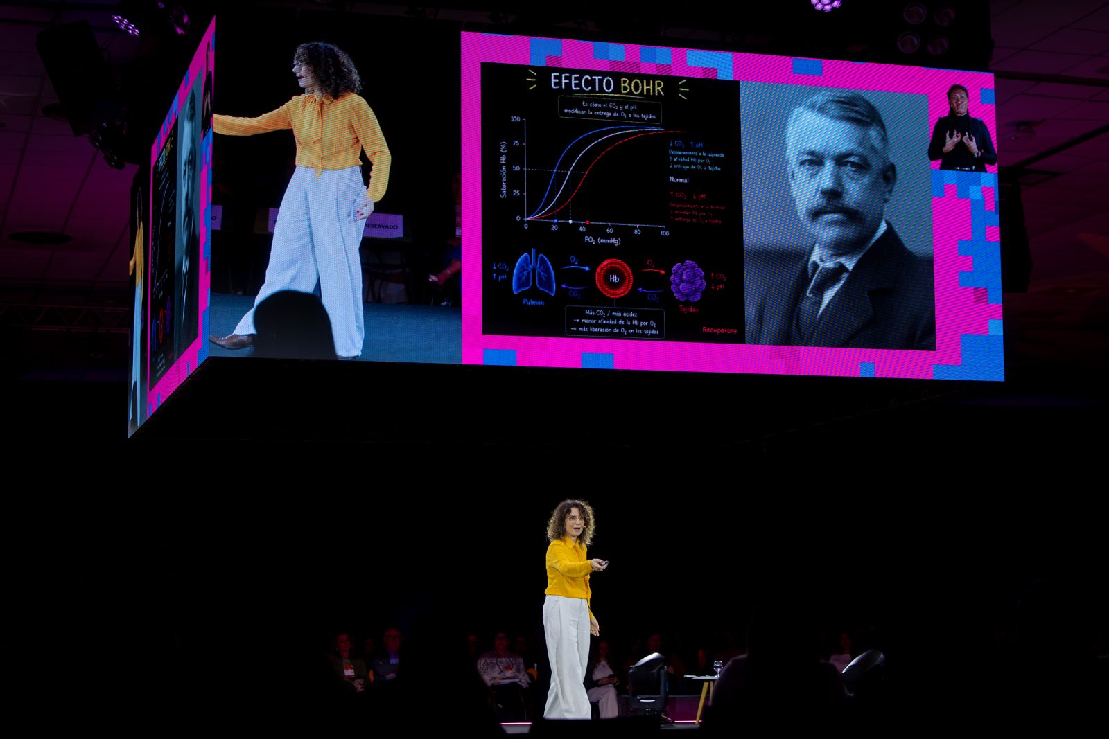 Fera Carvalho Leite no palco principal, sob o telão com um slide da palestra