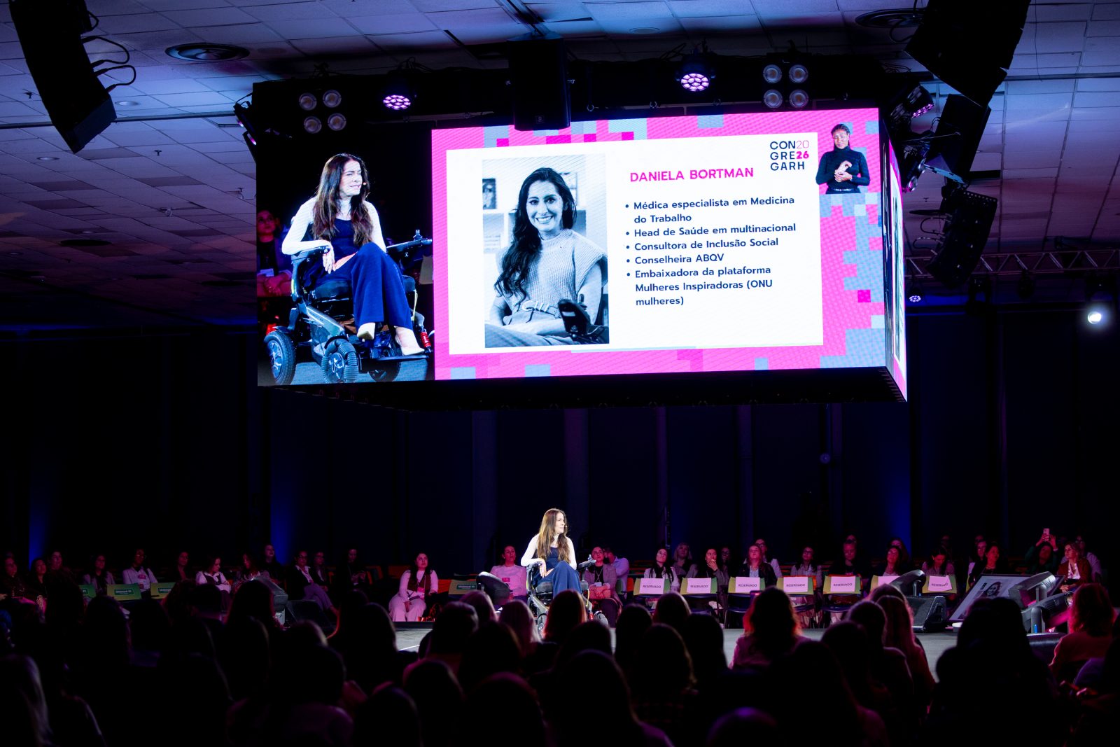 Dra. Daniela Bortman no palco principal, com o telão mostrando o slide de apresentação dela