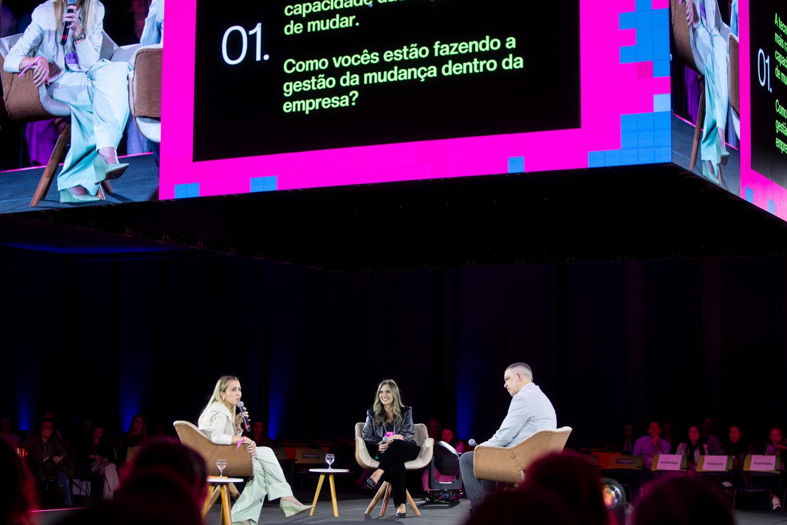 Carolina Ferreira, Fernanda Smith Bastian Brunetto e Rafael Martins no palco principal, sob o telão com uma pergunta do painel